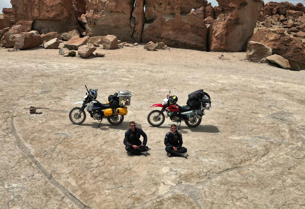 Zwei Motorradfahrer sitzen neben ihren Motorrädern auf steinigem Boden in Bolivien während Motorradtour durch Südamerika